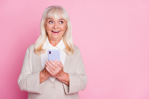 Photo portrait of excited old woman holding phone in two hands isolated on pastel pink colored background.