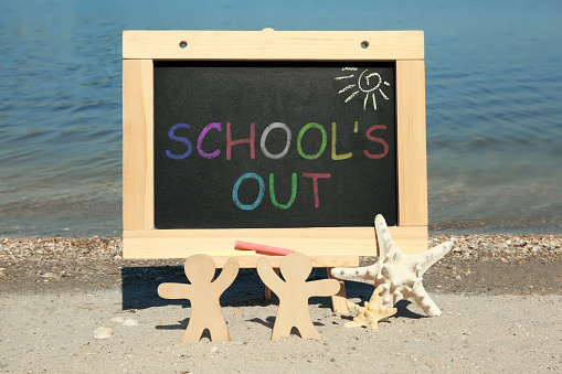 Chalkboard with text SCHOOL'S OUT, wooden people and starfish on beach