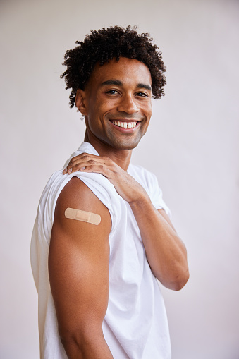 Portrait of a smiling young African man with a bandage on his arm after a Covid-19 vaccination standing on a gray background