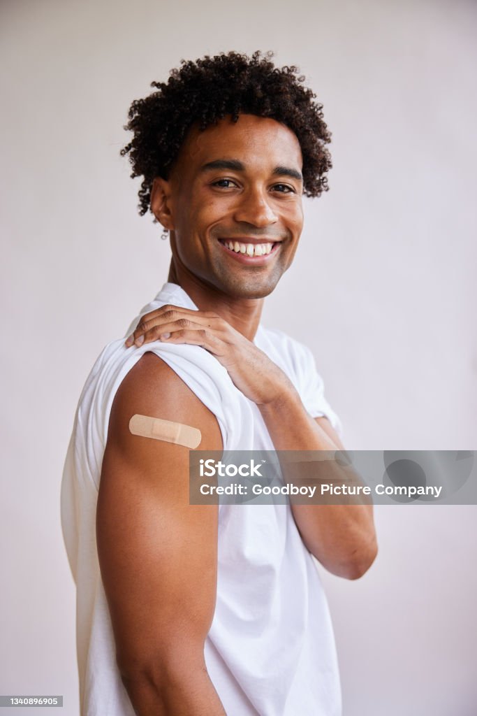 Smiling African American man with a bandage on his arm after a Covid-19 vaccination Portrait of a smiling young African man with a bandage on his arm after a Covid-19 vaccination standing on a gray background Vaccination Stock Photo