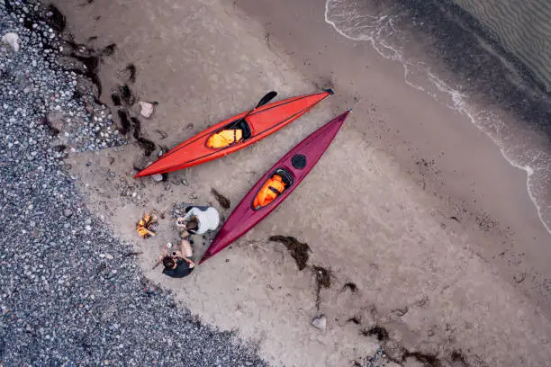 Drone’s eye view of a young couple making coffee by a campfire after a kayak trip. They are relaxing on the water’s edge with their sea kayak’s. Photographed on location on the island of Moen in Denmark. Colour, horizontal with some copy space.