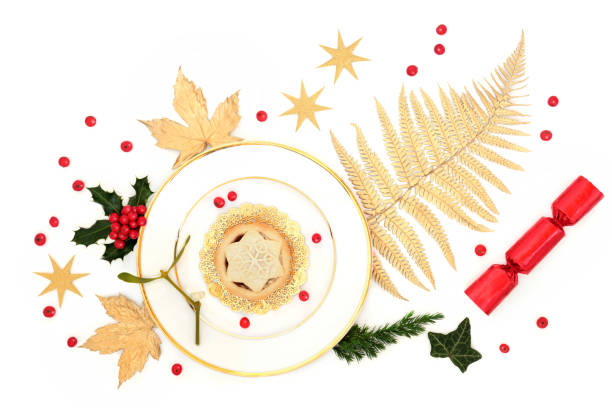 Luxury Christmas mince pie on a plate with gold leaves, stars, winter greenery, cracker and loose holly berries on white background.