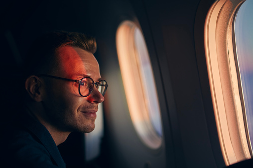 Young man with eyeglasses traveling by airplane. Passenger looking through window during flight at sunset.