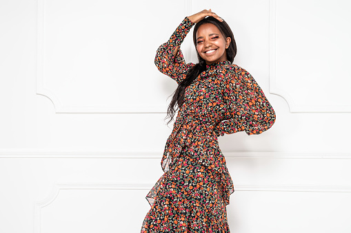Fashionable african american woman wearing floral pattern dress with long sleeves, posing on white wall, smiling and looking at the camera. Real people emotions and lifestyle.