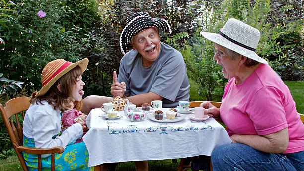 Grandparents and beautiful grandaughter having fun at a tea party Grandparents and 6 year old granddaughter are sitting at a small table in a garden, having a tea party and making funny faces at each other. Straw hats, china tea cups, cupcakes, and embroidered tablecloth are accessories. Horizontal format. antique chinese dolls pictures stock pictures, royalty-free photos & images