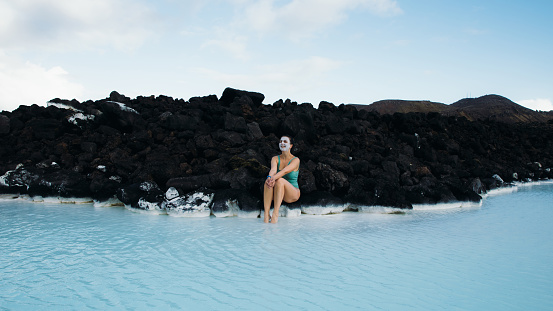 Woman traveler covered by white mud face mask relaxing in the pure blue hot spring thermal pool enjoying a journey in Iceland