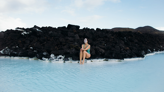 Woman traveler covered by white mud face mask relaxing in the pure blue hot spring thermal pool enjoying a journey in Iceland