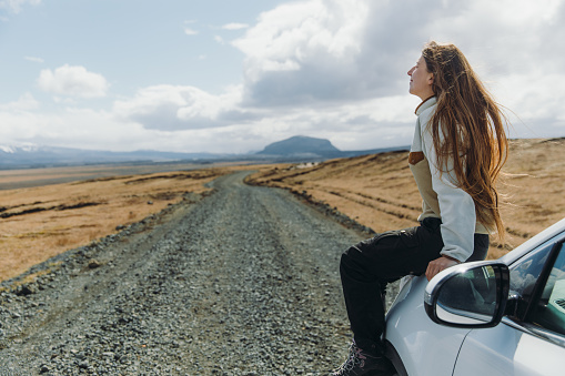 Smiling woman with long hair enjoying the road trip by the white car through the mountain landscape of Iceland