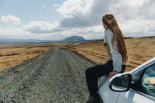 Smiling woman with long hair enjoying the road trip by the white car through the mountain landscape of Iceland