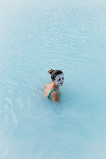 Woman traveler covered by white mud face mask relaxing in the pure blue hot spring thermal pool enjoying a journey in Iceland