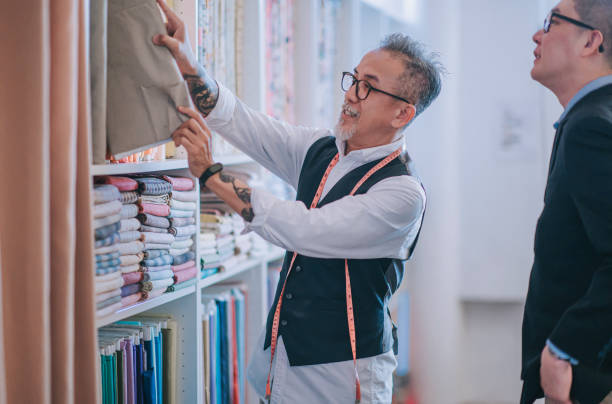 asiatico cinese senior uomo sarto che raccoglie e mostra al suo cliente la scelta del capo in tessuto su un nuovo suggerimento di abbigliamento su misura nel suo negozio - negozio di tessuti foto e immagini stock
