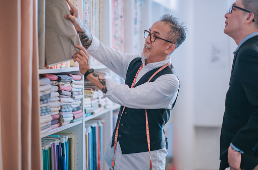 asian chinese senior man tailor picking and showing his customer on fabric garment choice on new tailored clothing suggestion at his store shelf display