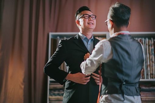 asian chinese senior man tailor helping his client trying on new tailored blazer suit at his studio