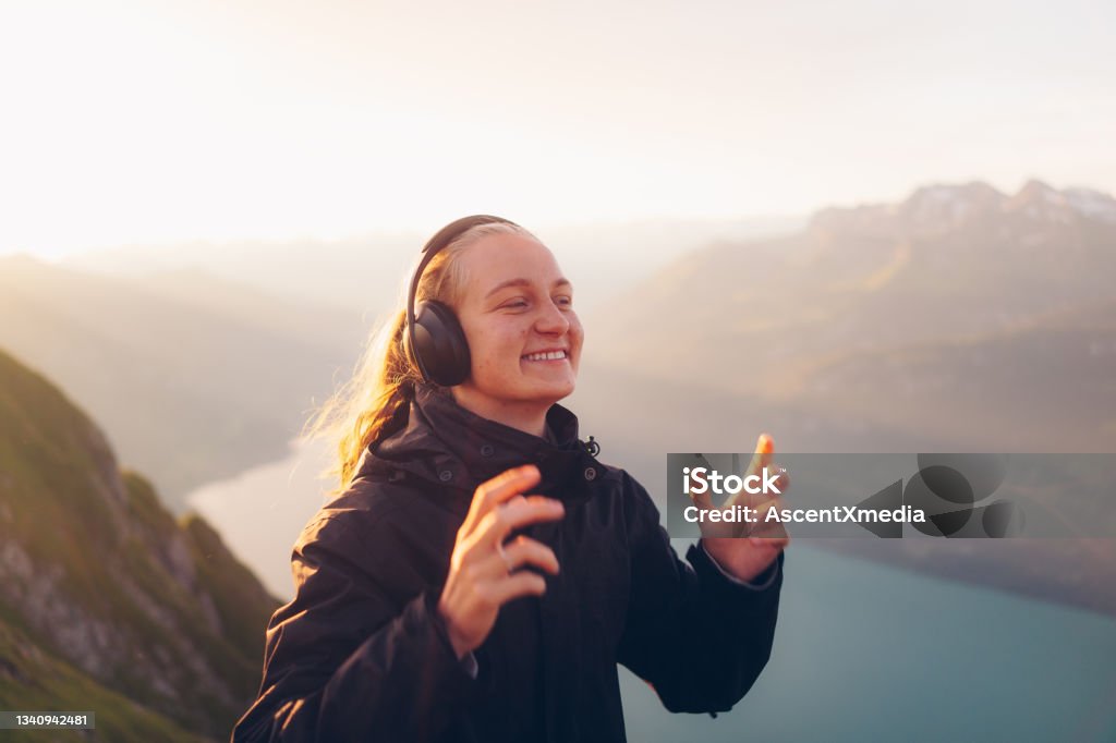 Female hiker listens to music at sunrise Lake Lugano visible below distant European Alps Listening Stock Photo