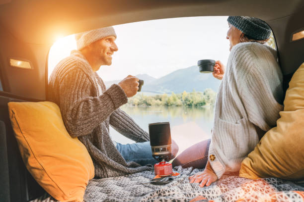 Car trunk view of chatting couple dressed warm knitted clothes enjoying gas stove prepared coffee and mountain lake view. Cozy early autumn couple auto traveling concept image. vista del maletero del coche de la pareja charlando vestidos de punto abrigado disfrutando de la estufa de gas preparada café y vista al lago de la montaña. acogedora imagen de concepto de viaje en automóvil para parejas de principios de otoño.