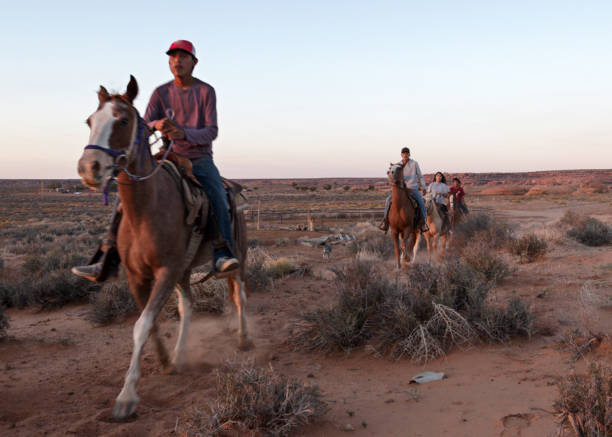 adolescenti e bambini navajo nativi americani che cavalcano cavalli insieme nella monument valley arizona, utah al crepuscolo sotto un tramonto drammatico alla fine dell'estate nel parco tribale - cherokee foto e immagini stock