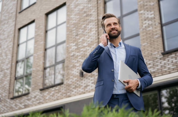 uomo d'affari che indossa un abito elegante che parla al cellulare, tenendo il laptop in piedi per strada. business di successo - outdoors business computer laptop foto e immagini stock