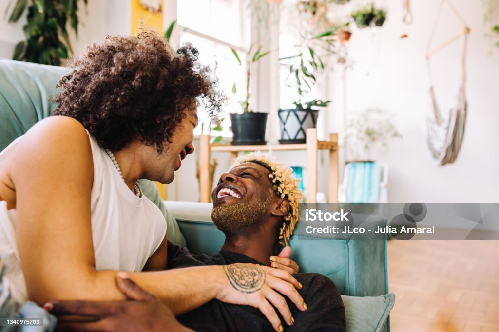 Cheerful young gay couple being romantic at home Cheerful young gay couple being romantic indoors. Two romantic young male lovers smiling at each other while lying on the couch in their living room. Happy young gay couple bonding at home. African-American Ethnicity Stock Photo