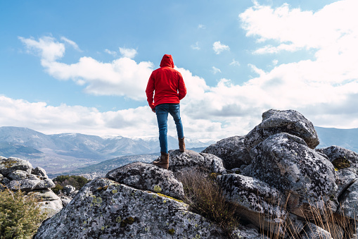 unrecognizable man standing dressed in red jacket on the rocks on top of a mountain looking at the mountains under a blue sky with clouds