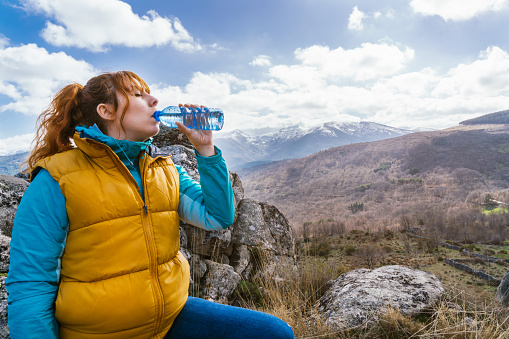 a red-haired girl in a yellow vest resting and drinking water sitting on a rock on top of a mountain on a sunny day. Gredos. Spain.