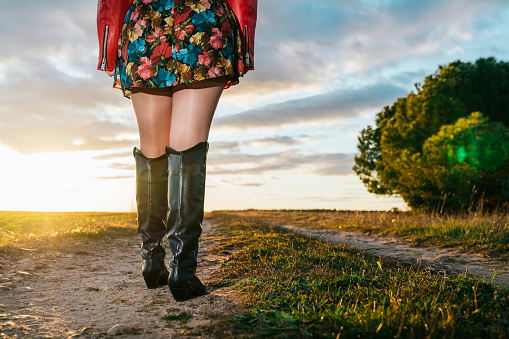 Closeup of a woman's legs wearing black cowboy boots walking in the field at sunset under a blue sky with clouds. copy space.