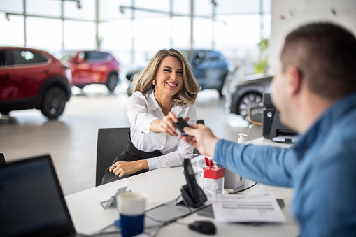 Salesman sitting with young female customer in car dealership and giving her car keys