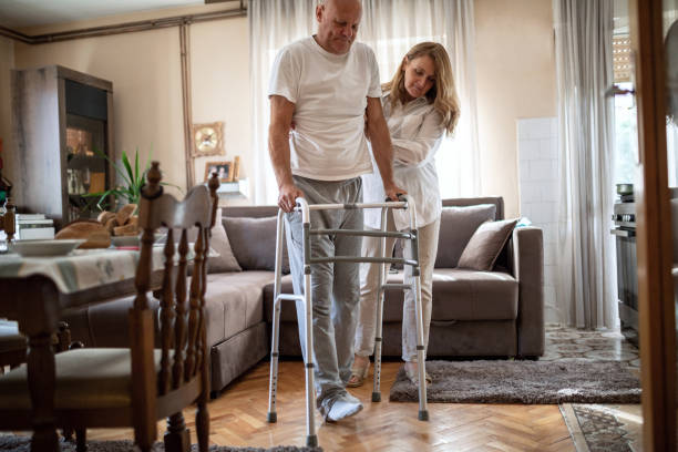Wife helping her injured husband to walk using walking frame at home stock photo