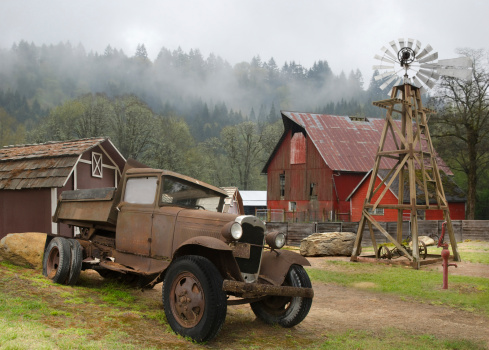 Old American farm featuring a rusty, vintage truck, red barns, windmill, water pump and misty hills in the background
