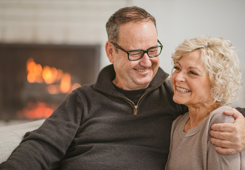 A senior couple sit together closely on a sofa in front of a roaring fireplace.  The husband has his arm around his wife pulling her in closely to hug her, while she smiles back at him with glee.