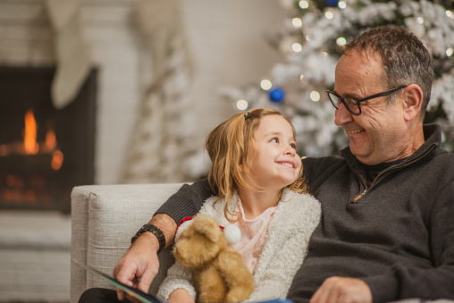A Grandfather sits with his Granddaughter snuggled in closely while on the sofa at Christmas.  They are both dressed casually in sweaters and looking at one another with a smile.