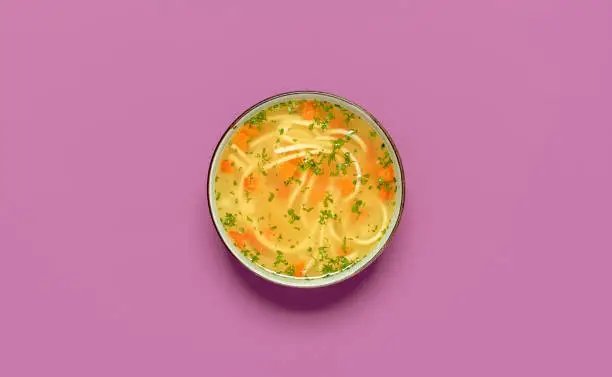Top view with a bowl of chicken soup isolated on a purple background. Delicious homemade chicken soup with vegetables and noodles.
