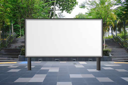 Large blank advertising poster billboard banner mockup in front of stairs; lush plants and tress in background; digital light box display screen for OOH media. Speckled textured concrete cement ground