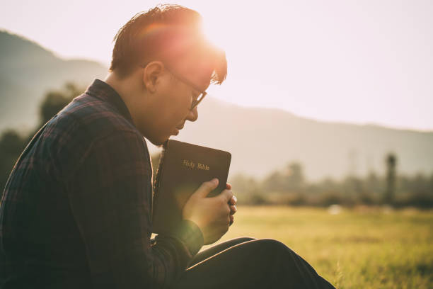 hombre orando en la santa biblia en un campo durante la hermosa puesta de sol.hombre sentado con los ojos cerrados con la biblia en sus manos, concepto de fe, espiritualidad y religión. - biblia fotografías e imágenes de stock