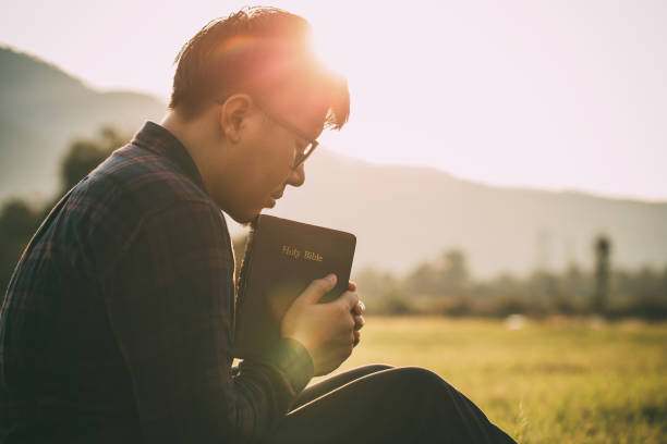 man praying on the holy bible in a field during beautiful sunset.male sitting with closed eyes with the bible in his hands, concept for faith, spirituality, and religion. - bijbel stockfoto's en -beelden