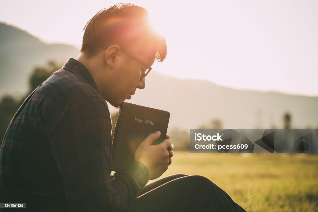 man praying on the holy bible in a field during beautiful sunset.male sitting with closed eyes with the Bible in his hands, Concept for faith, spirituality, and religion. Praying Stock Photo