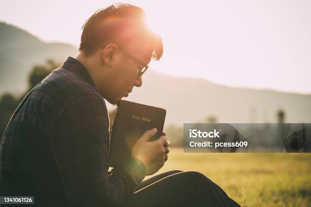 Man Praying On The Holy Bible In A Field During Beautiful Sunsetmale Sitting With Closed Eyes With The Bible In His Hands Concept For Faith Spirituality And Religion Stock Photo - Download Image Now