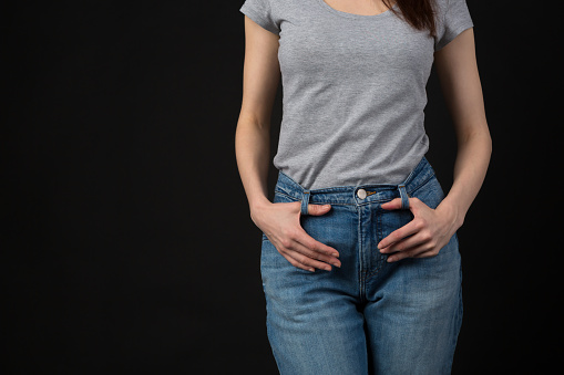 Slim young woman wearing classic blue jeans and casual grey shirt in studio. Unrecognizable female model body part with hands on her waist