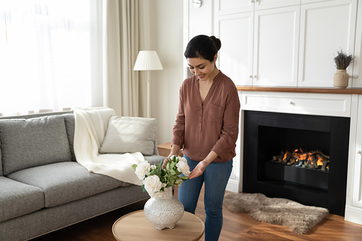 Cozy homey. Happy millennial indian woman young wife decorating luxury living room of family home put bunch of white roses on table before fireplace. Young mixed race female improving house interior