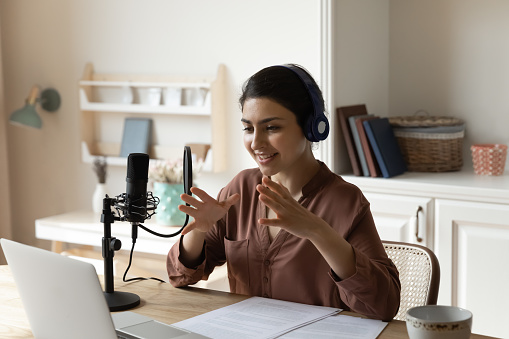 Broadcasting online. Artistic indian woman in headphones sit by desk with laptop speak using professional microphone record audio podcast. Young lady business coach shoot video training at home studio
