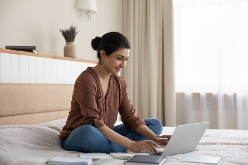 Preparing for exam. Confident millennial mixed race woman sit on bed engaged in distant studying using laptop pc. Motivated indian female student do research homework search learning material online