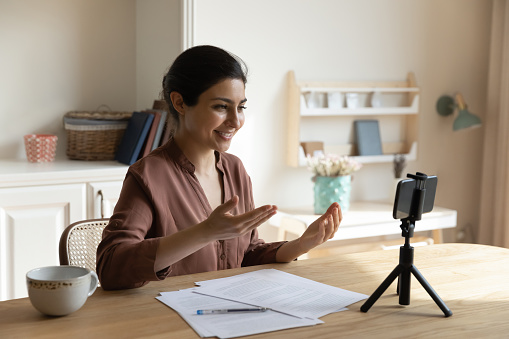 Videoblogging. Happy young lady vlogger of indian ethnicity sit at desk before smartphone on holder talk to audience record video review. Mixed race female teacher shoot lecture course on phone webcam