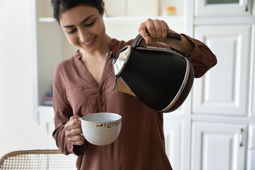 Making tea. Happy young woman of indian ethnicity hold cup teapot in hands brew instant coffee black green herbal tea to drink with pleasure. Millennial hindu female prepare hot drink in ceramic mug