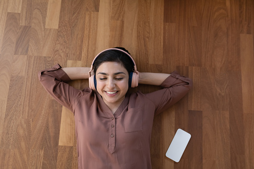 Music break. Top view of restful indian female in modern headset lie on heated warm floor listen to songs on smartphone. Young lady enjoy audio tracks from cell playlist with closed eyes. Copy space
