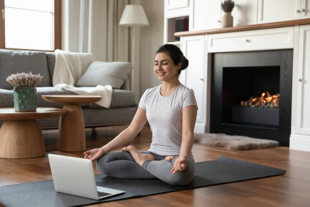 giovane donna indiana sportiva medita sul tappeto prima del moderno laptop - yoga class instructor yoga exercising foto e immagini stock