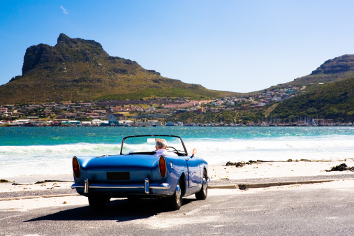 Elderly man sits in his vintage British sports car, watching the sea and a fishing harbor across the bay.