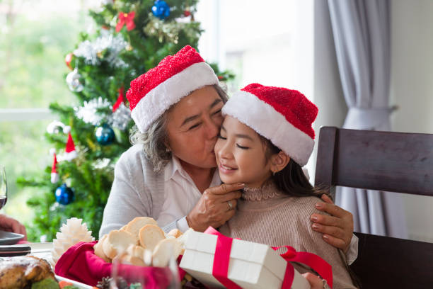 счастливая азиатская семья на рождество. бабушка целует милую маленькую девочку, держа в руках рождественскую подарочную коробку и сидя, о� - senior adult winter senior women daughter стоковые фото и изображения
