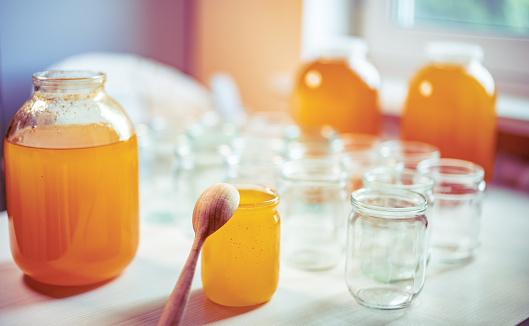 Composition of large number of empty glass jar and three large glass transparent jar with yellow sweet honey and small wooden spoon, standing on large white table against background of bright sunlight