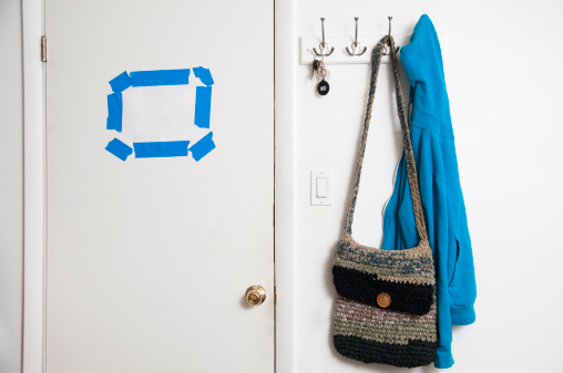 Blank note stuck with blue tape to a white door. A key ring, a purse, and a blue jacket hanging on the coat hanger nearby. Copy space.