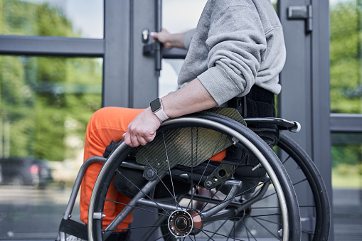 Cropped view of the physically challenged girl moving herself at the wheelchair and opening doors of the building. Stock photo