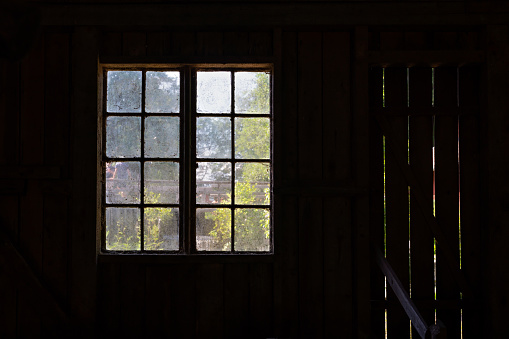 Looking through an old dirty farm house window from the inside with summer view. Dark interior with worn wooden walls I Vira Bruk Stockholm county Sweden.